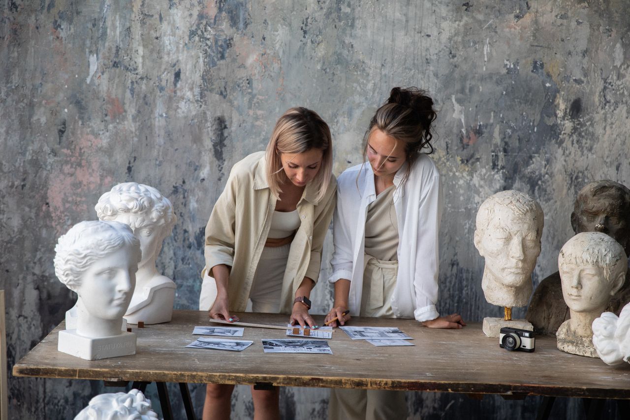 Two women study sculpture references in an art studio with sculpted busts.